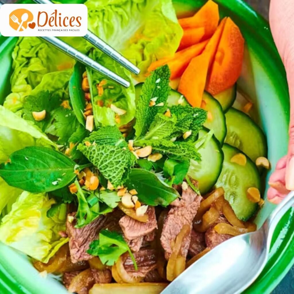 Gros plan sur un bol de Bo Bun Bœuf très frais, avec des morceaux de bœuf, des vermicelles de riz, des concombres, des carottes et de la menthe. Une main tient des baguettes. Image de la recette bo bun bœuf traditionnelle.