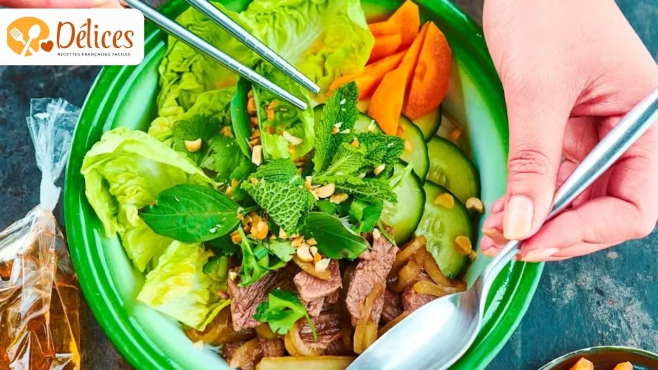 Gros plan sur un bol de Bo Bun Bœuf très frais, avec des morceaux de bœuf, des vermicelles de riz, des concombres, de la menthe et de la salade. Une main tient des baguettes. Image de la recette bo bun boeuf.